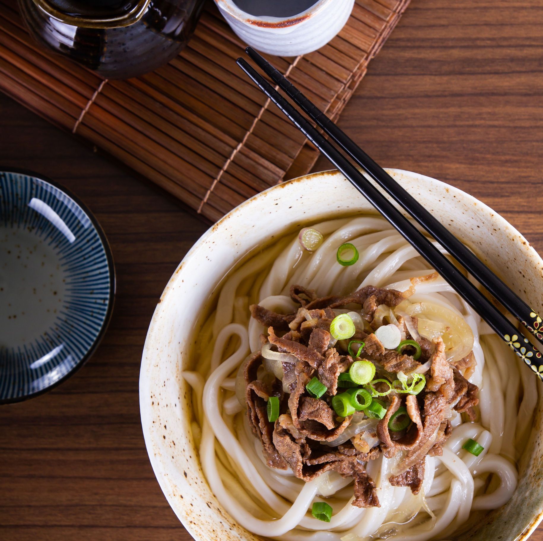 Bowl of udon noodles topped with beef and green onions, with chopsticks on a wooden table.