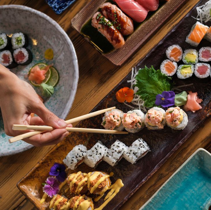 Hands using chopsticks over a sushi platter with assorted rolls and sashimi on wooden trays