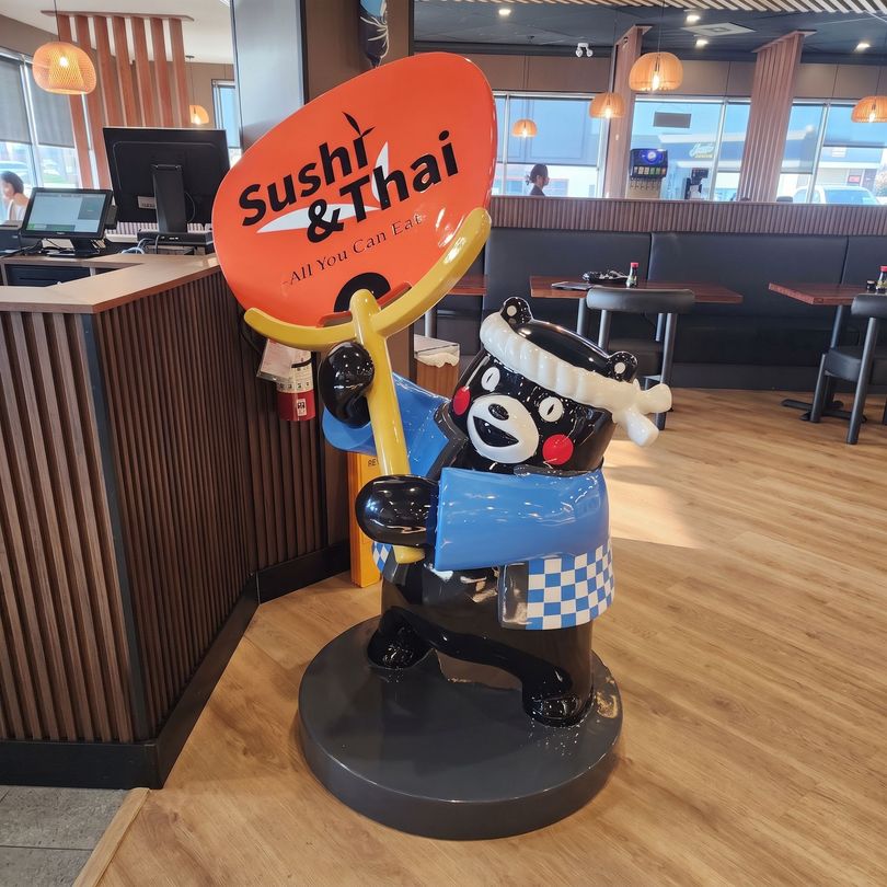 Kumamon mascot statue holding a Sushi Thai sign inside a restaurant, with wood floors and booths.
