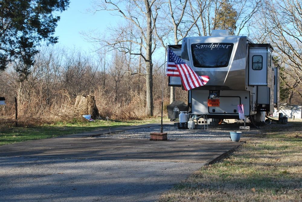RV parked at a campsite; American flag; trees in background.