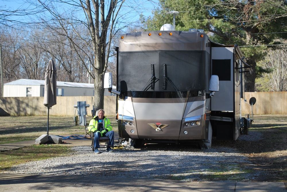 RV parked in a gravel lot with a person sitting outside. Trees and a small building in the background.