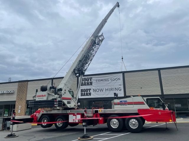 A large crane lifting a sign for Boot Barn at a shopping center. The crane is red and white. Cloudy sky.