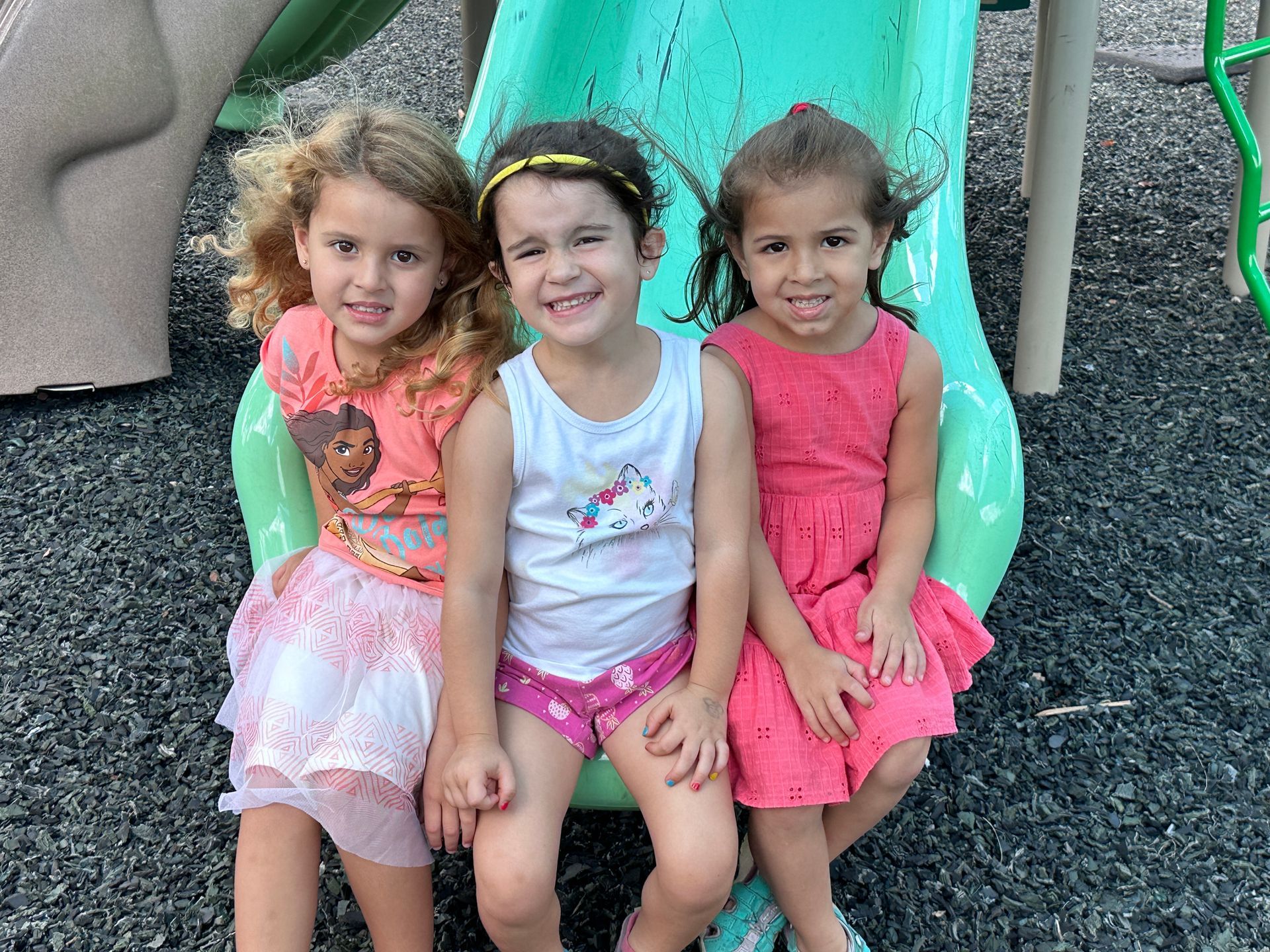 Three children sit smiling together on a green playground slide outdoors.