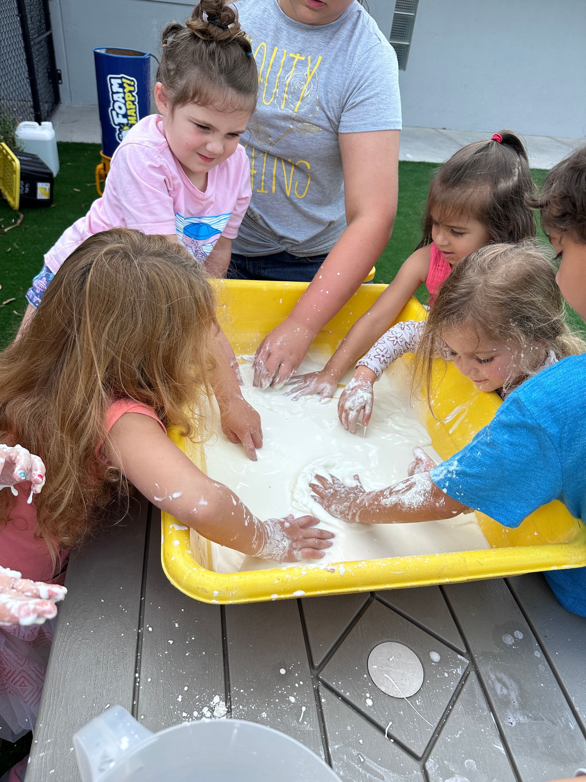 Children gather around a yellow bin to play with a white, powdery substance spread across a table.