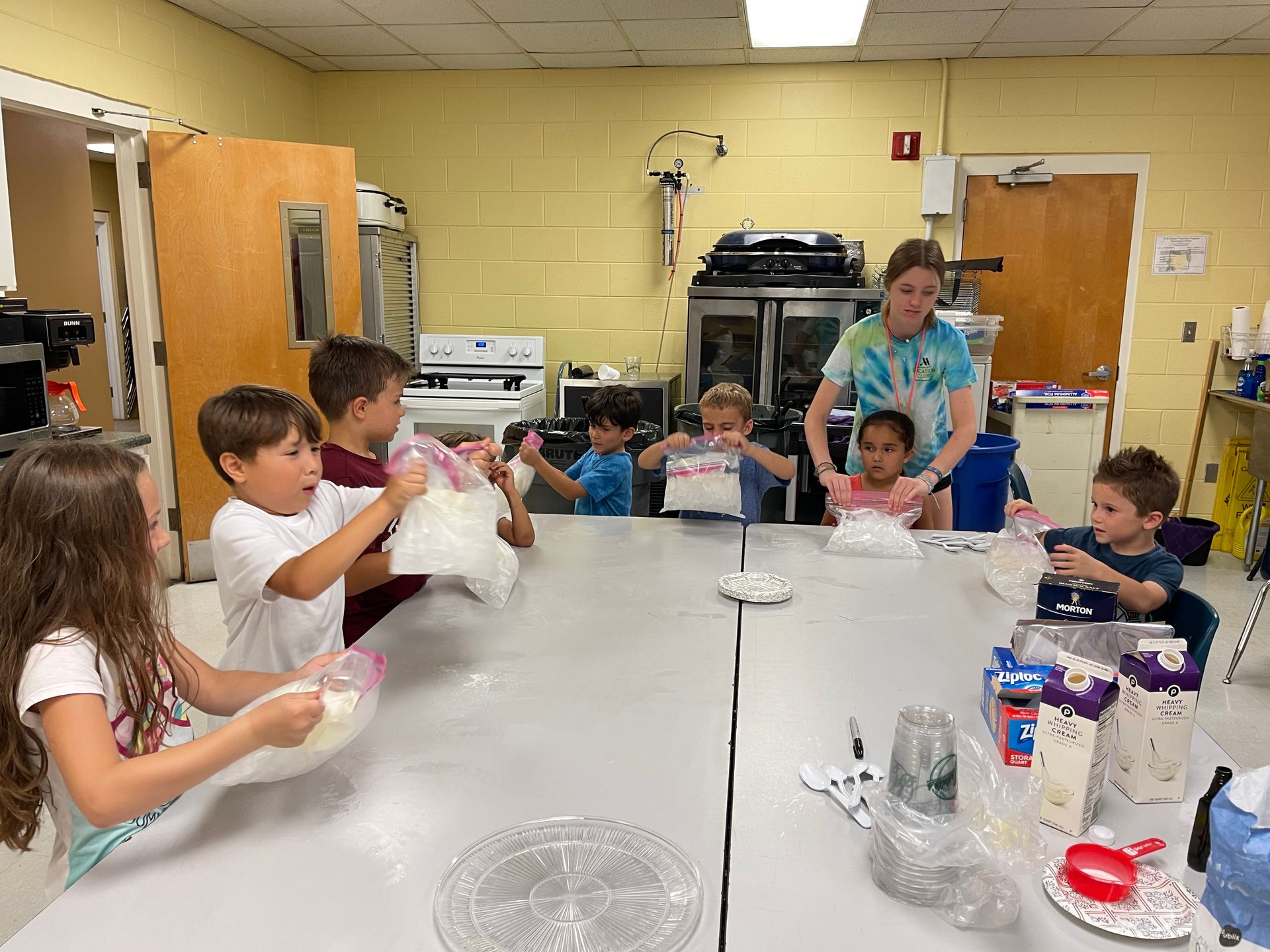 A group of children and an adult at a large table inside a kitchen, filling plastic bags with ingredients for an activity.