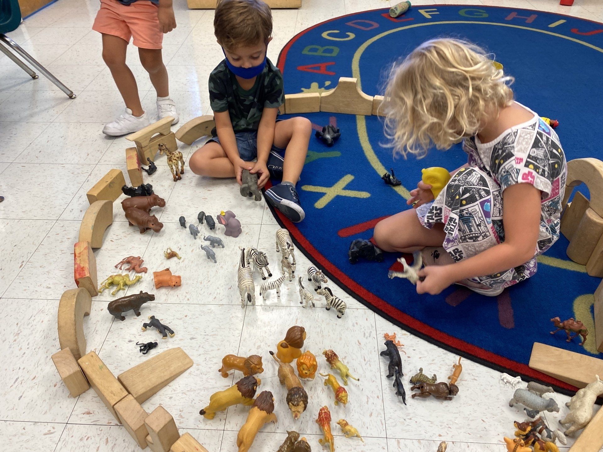 Two children sit on a rug playing with many small toy animals inside a wooden block enclosure in a classroom.