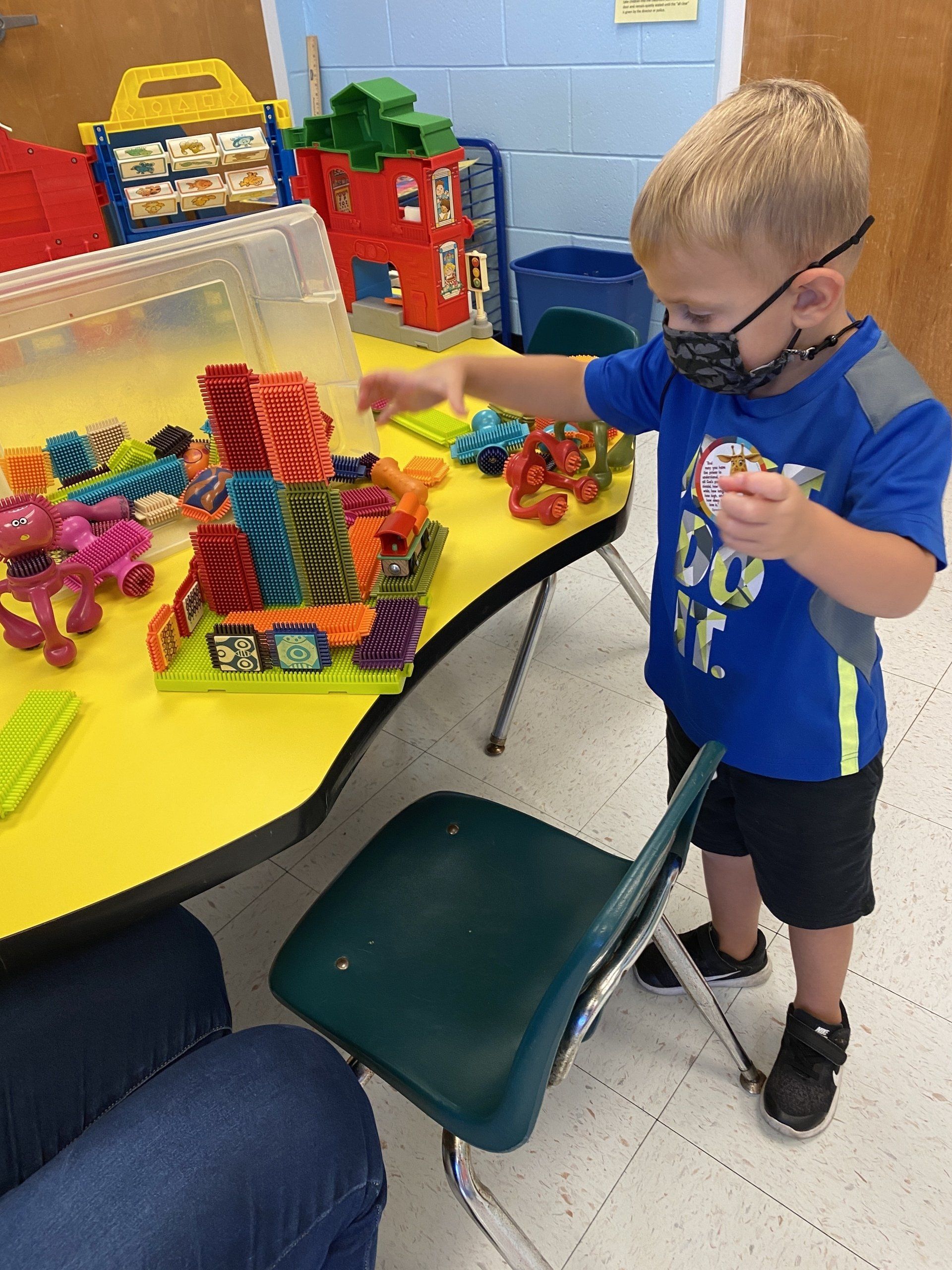 A child wearing a mask stands at a yellow table in a classroom, building a structure with colorful plastic connectors.