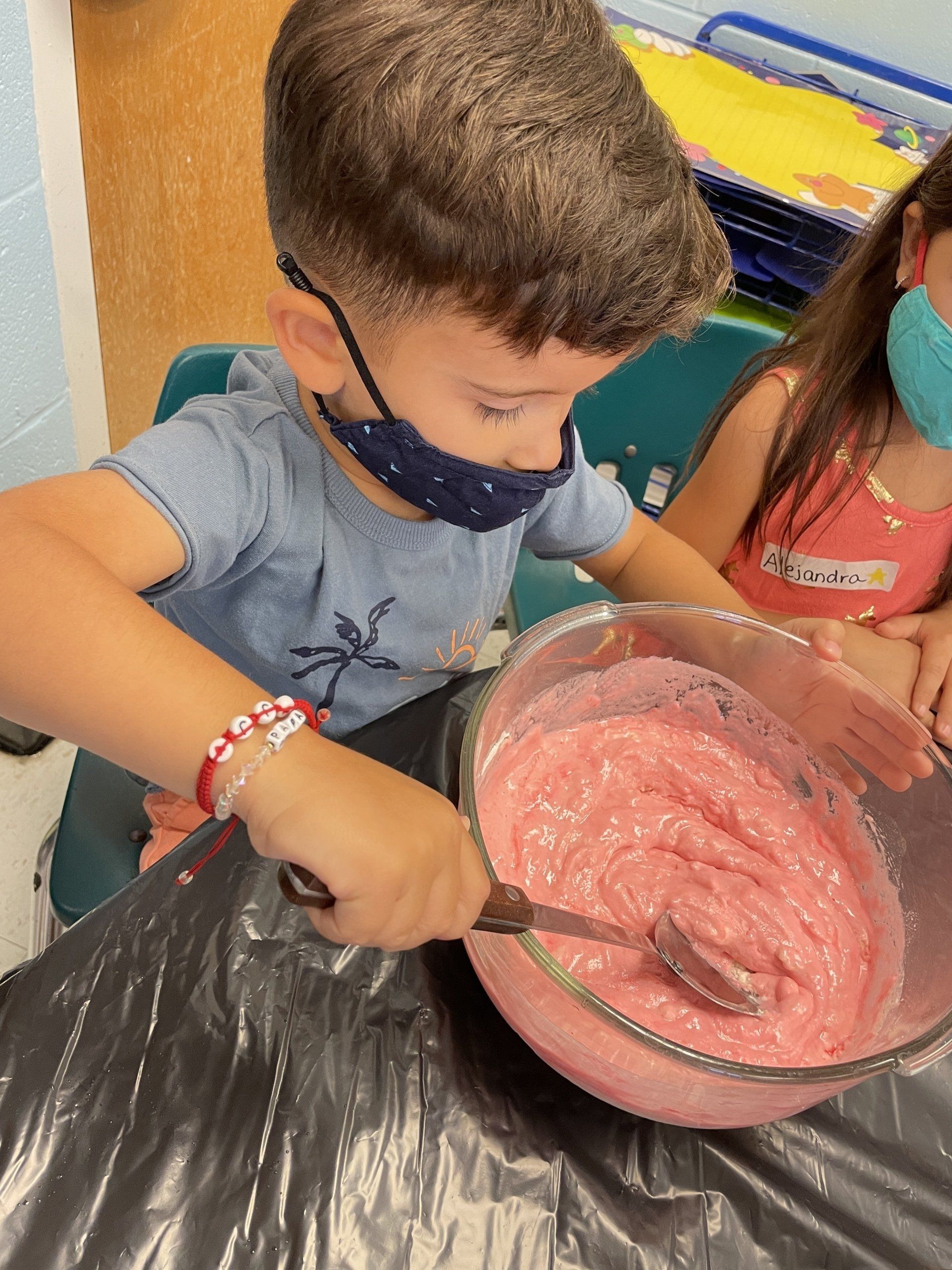 A child wearing a mask stirs pink batter in a clear bowl while sitting at a table.
