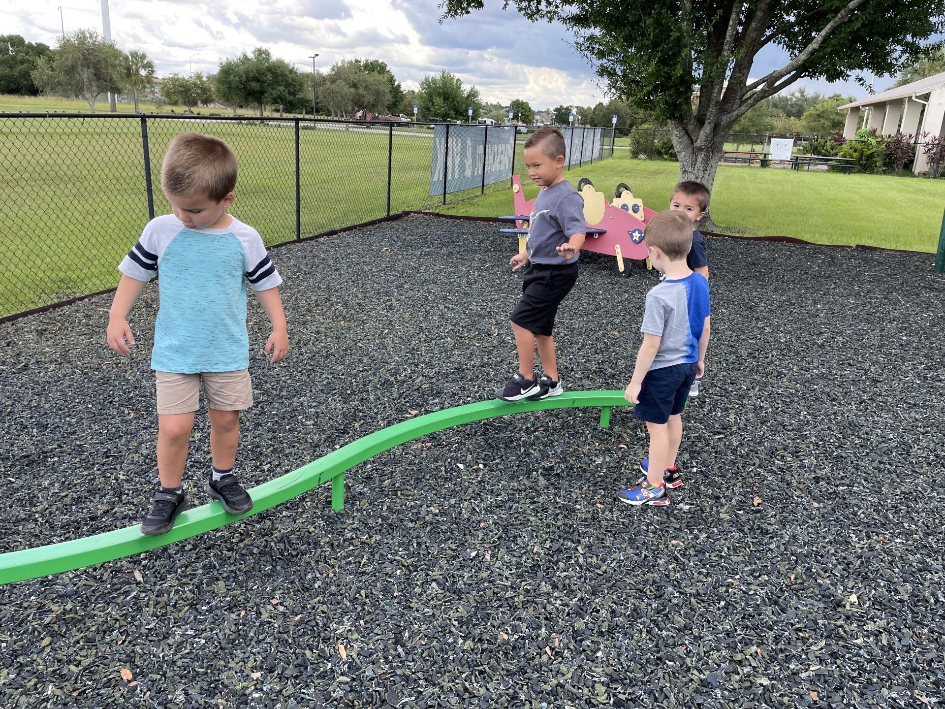 Four children balance on a low, green curved beam in a gravel playground.