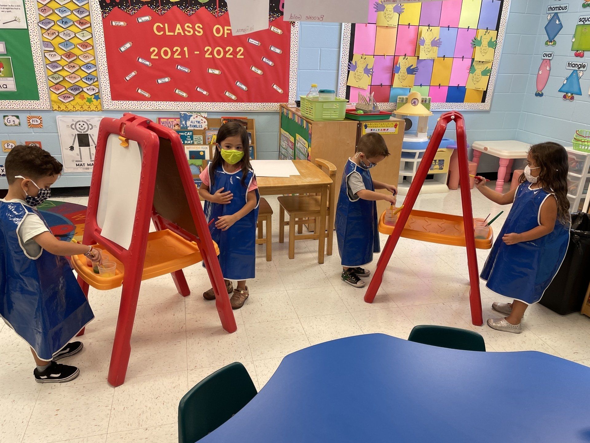 Four children wearing blue art smocks stand at two red easels inside a brightly decorated classroom.