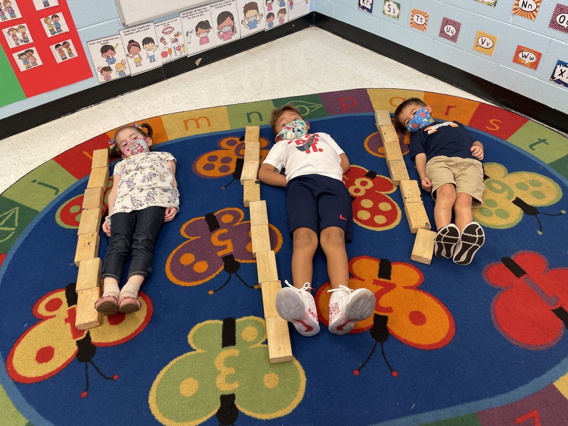 Three children lie on their backs on a butterfly-patterned rug, each flanked by lines of wooden building blocks.