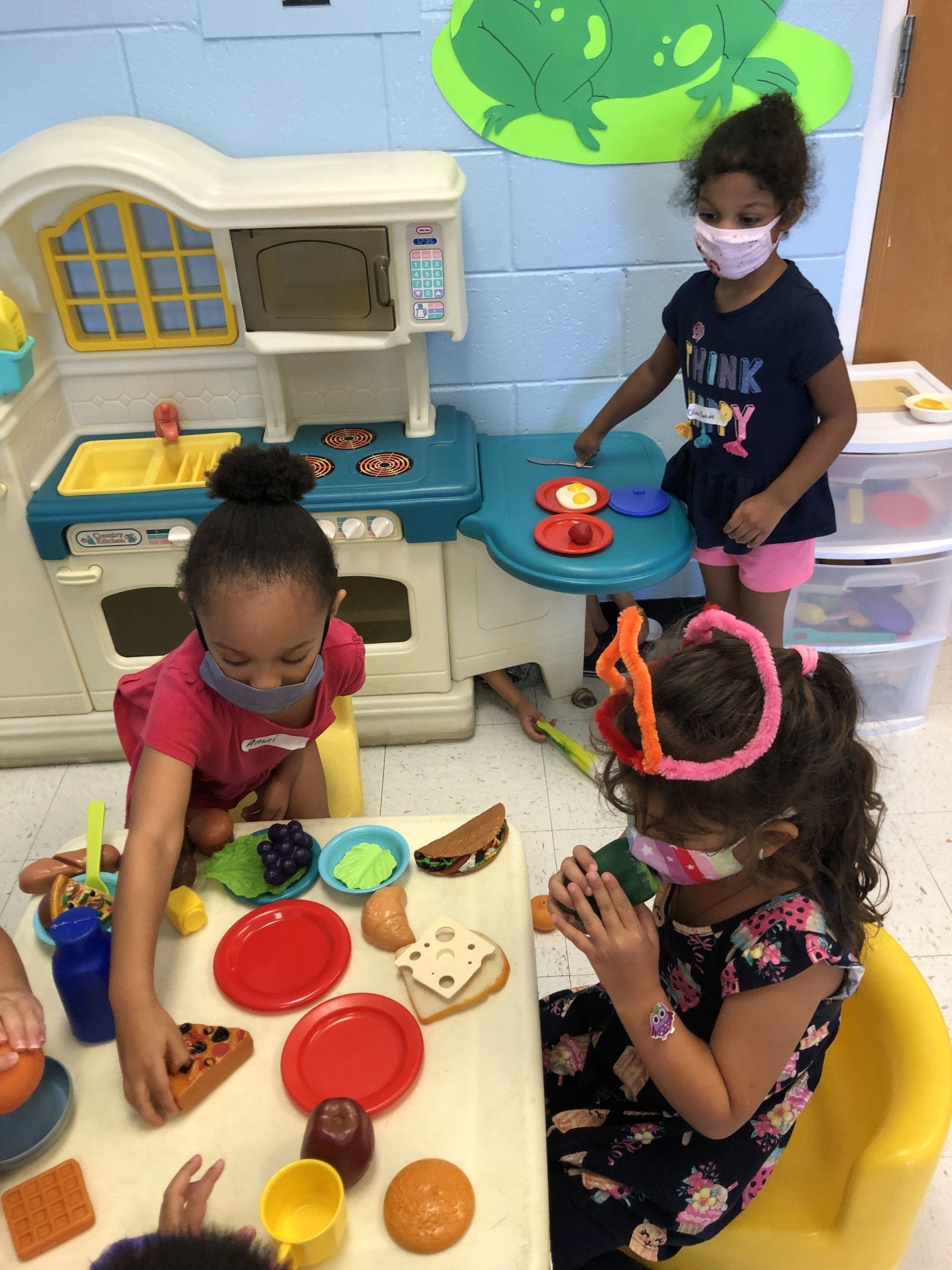 Three children wearing face masks play together in a colorful classroom, using a toy kitchen and setting a plastic table.