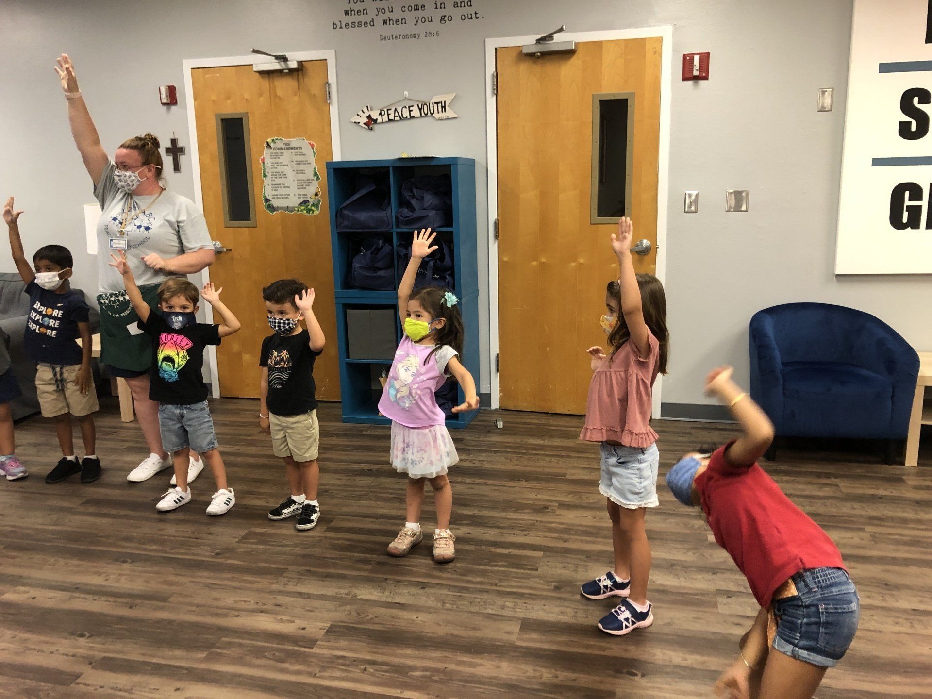 A group of children and an adult in a classroom stand and raise their hands during an activity.