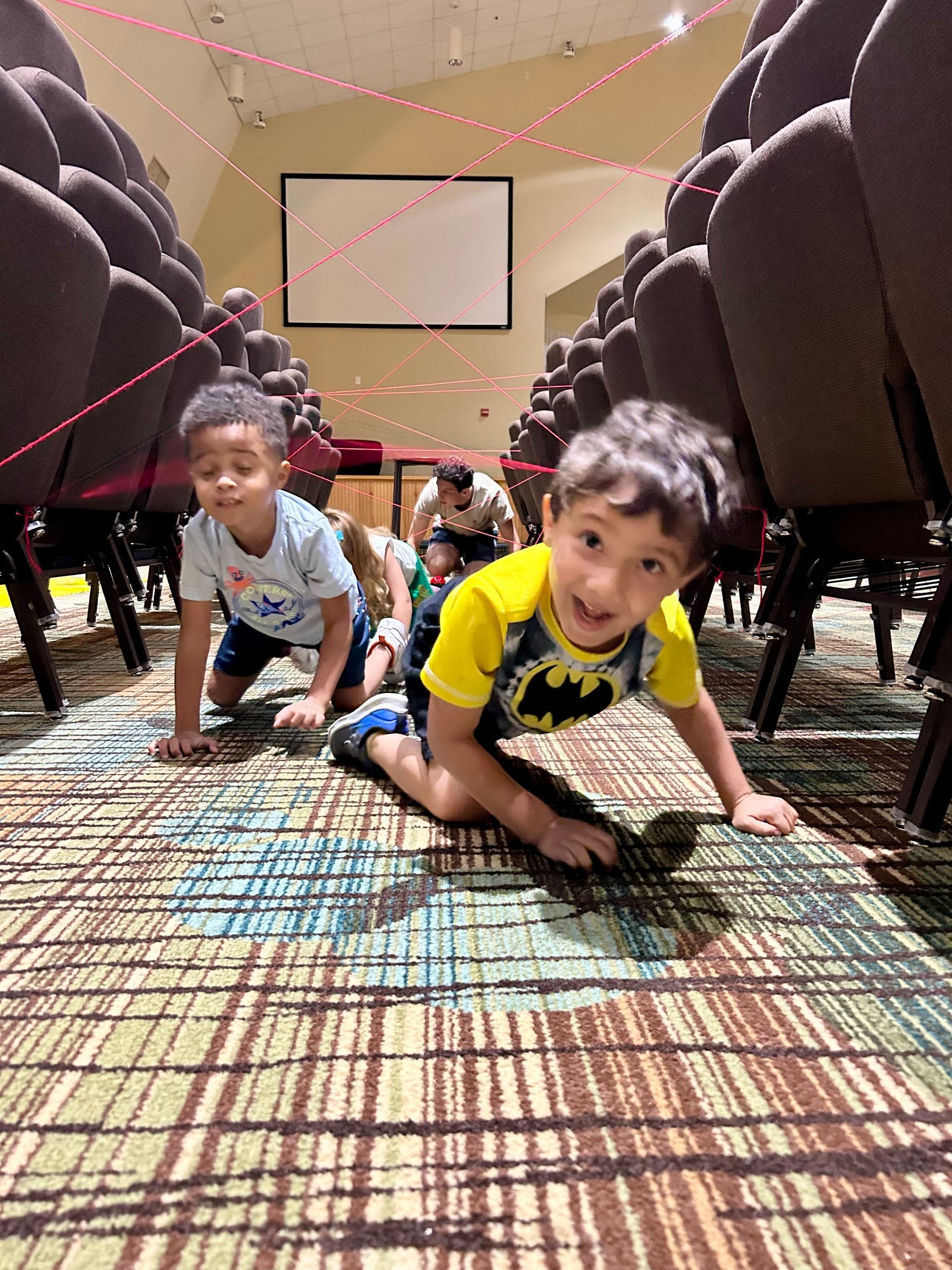 Two children crawl on a carpeted floor between rows of theater seats, navigating a course made of red string.