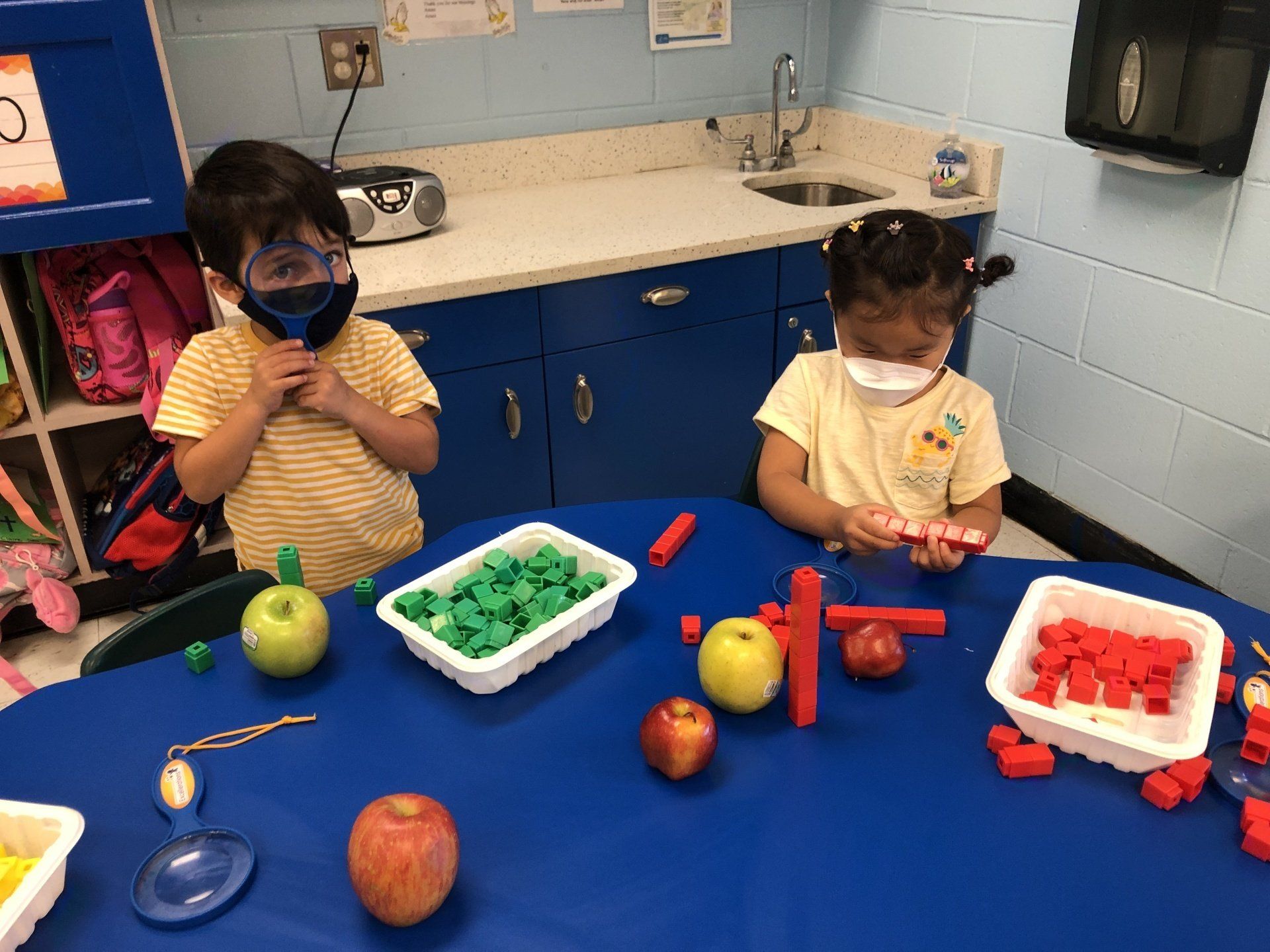Two children wearing masks at a table with apples, plastic fruit, and trays of red and green connecting blocks.
