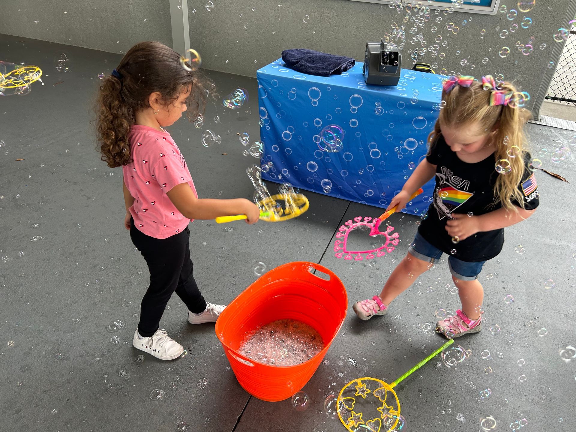 Two children play with bubble wands and solution in a bucket outdoors, surrounded by floating bubbles.