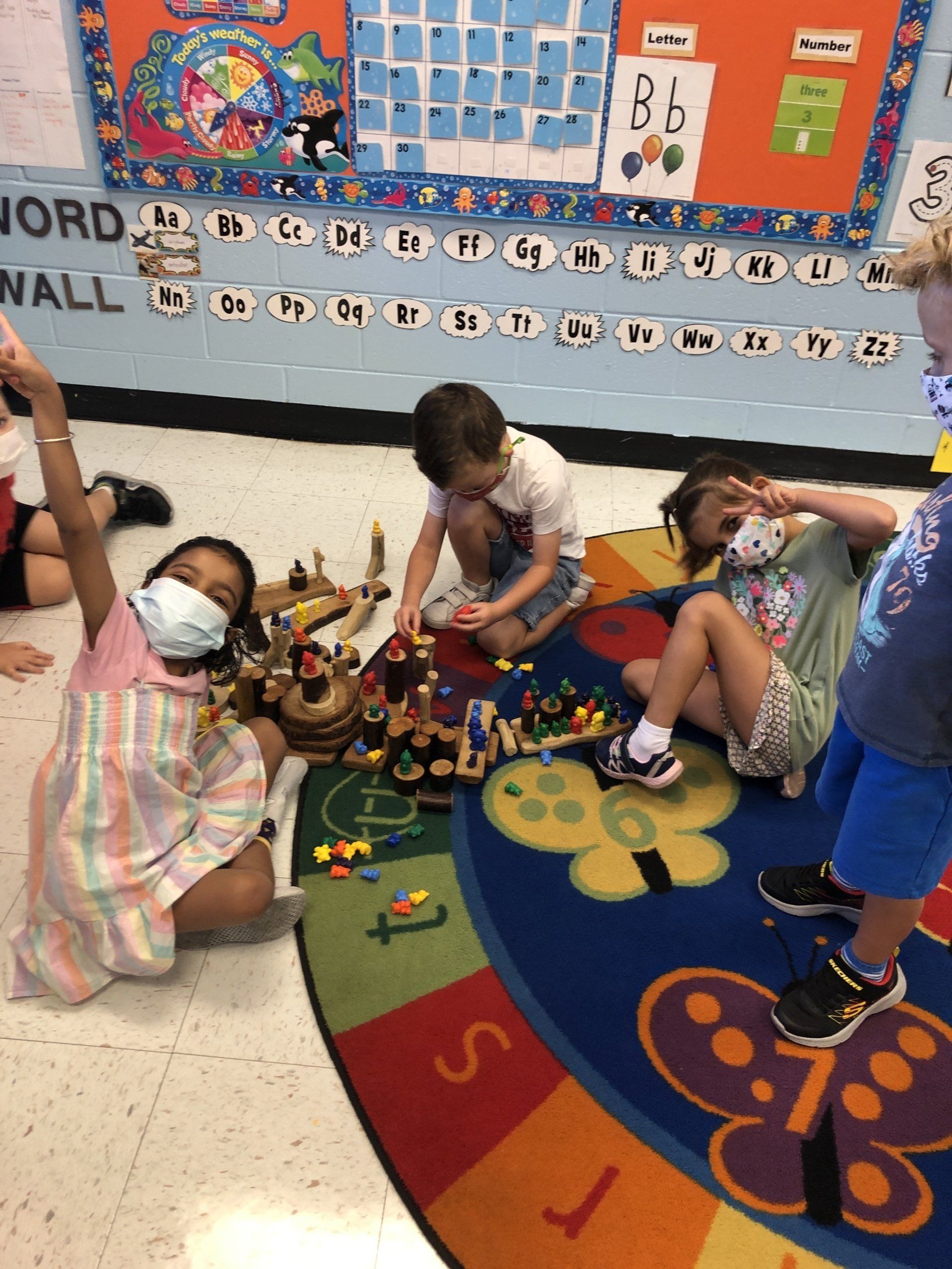 Children wearing face masks play with small toys on a colorful alphabet rug in a classroom.