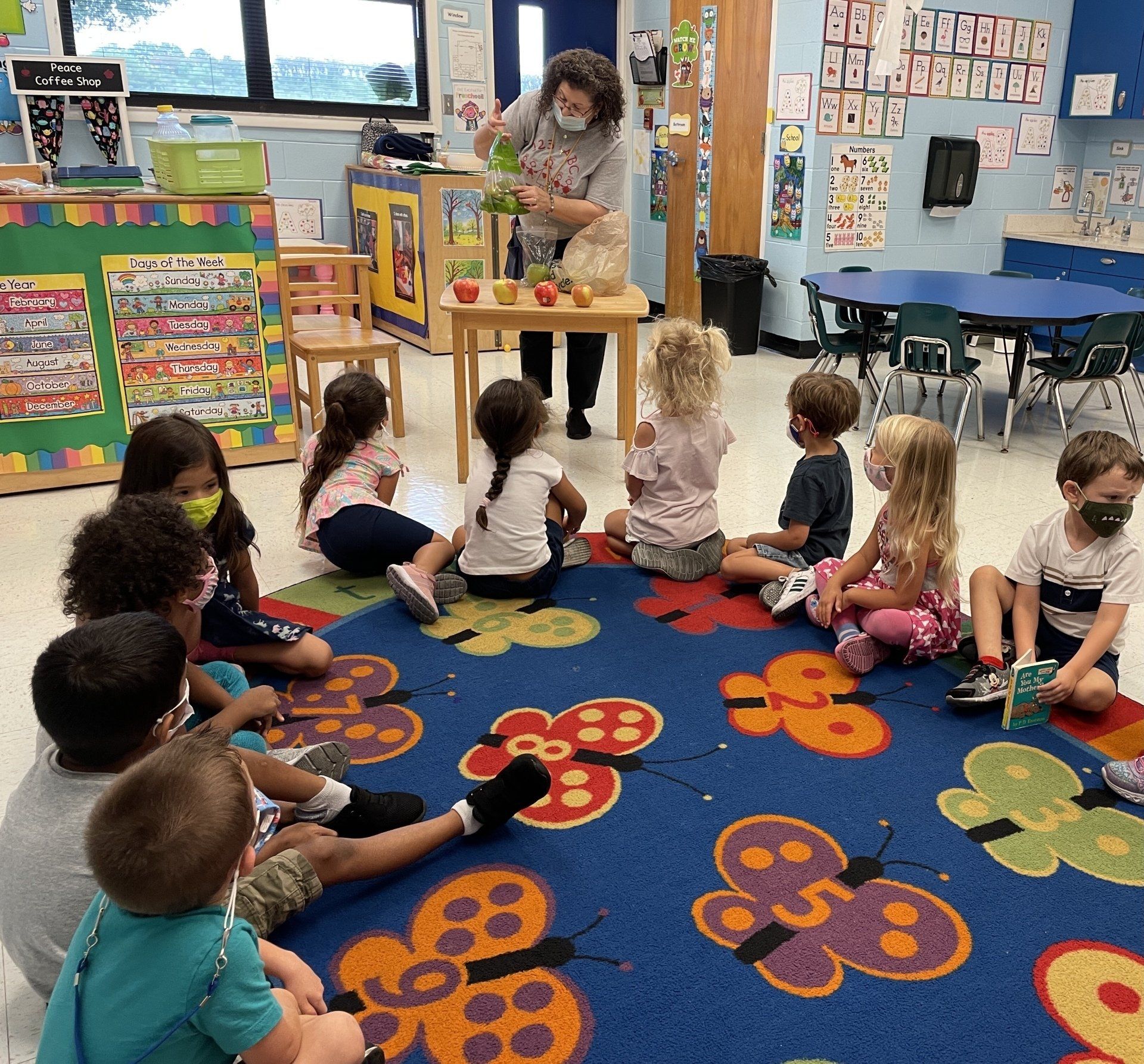 A teacher shows apples to a group of young students sitting on a colorful, numbered butterfly rug in a classroom.
