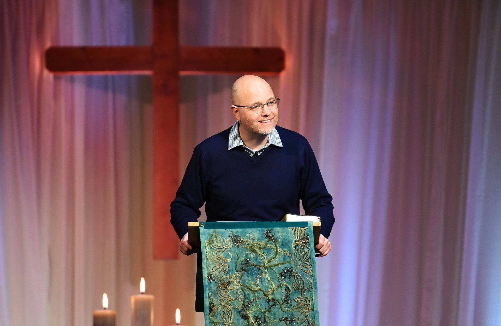 A person speaking at a podium with a decorated cover, standing in front of a cross and draped fabric in a church.