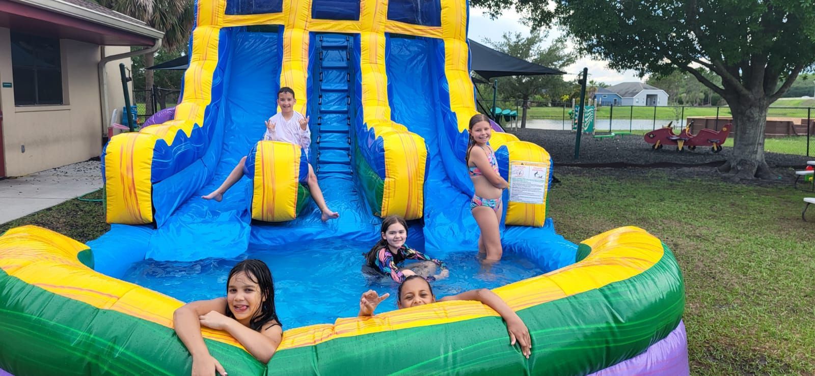 Four children playing in a blue, yellow, and green inflatable water slide in a grassy yard.