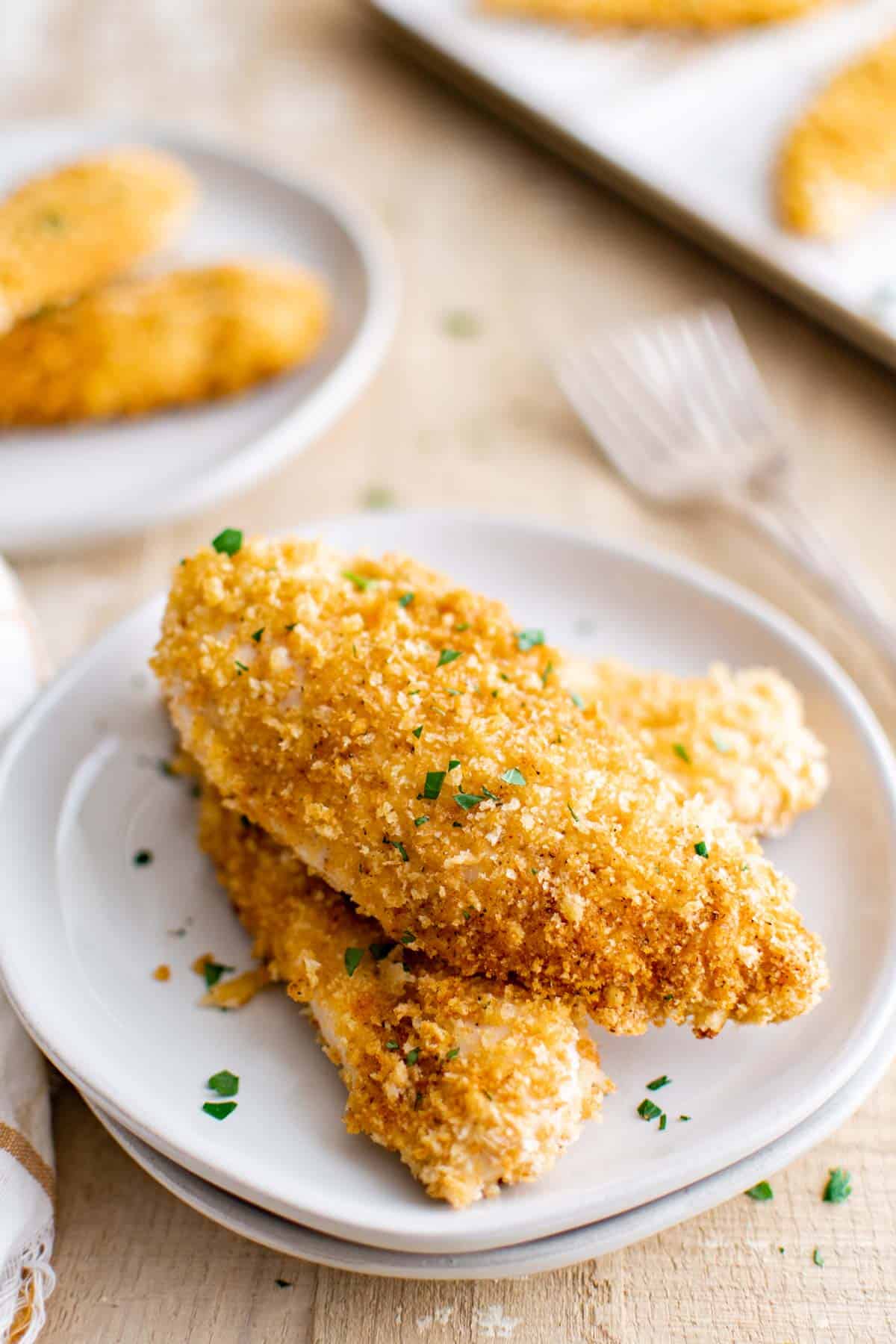 Golden, crispy breaded chicken tenders garnished with fresh green herbs, served on small white plates.