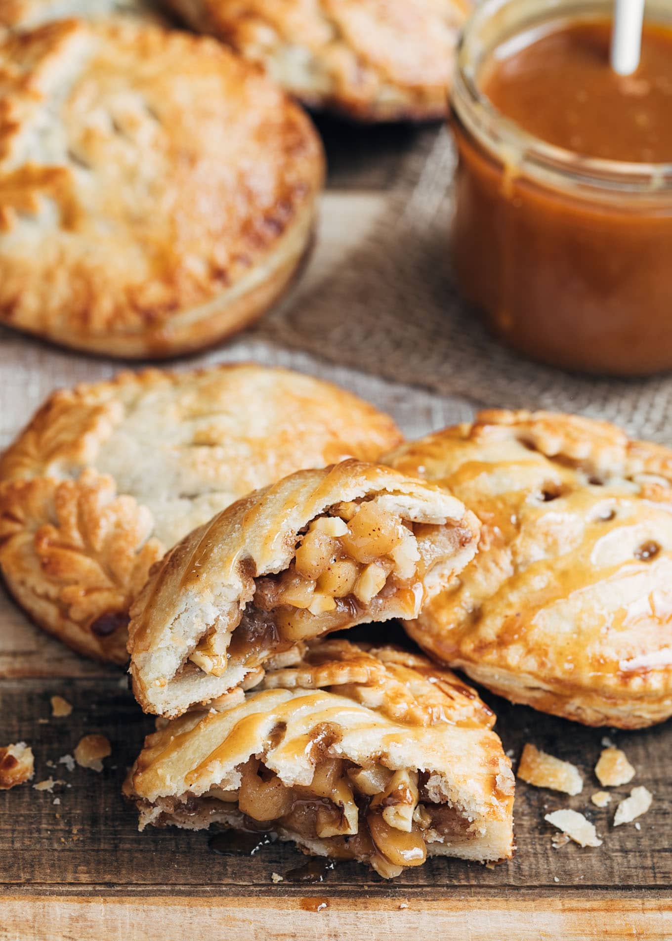 Several golden-brown apple hand pies, with one cut in half to show the filling, beside a jar of caramel sauce.