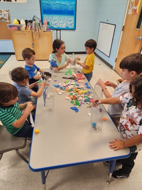 A group of students sitting around a table in a classroom sorting colorful craft materials into clear plastic bottles.