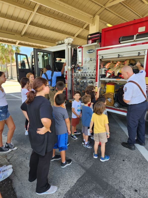 A firefighter talks to a group of children and adults near an open fire truck compartment at a fire station.