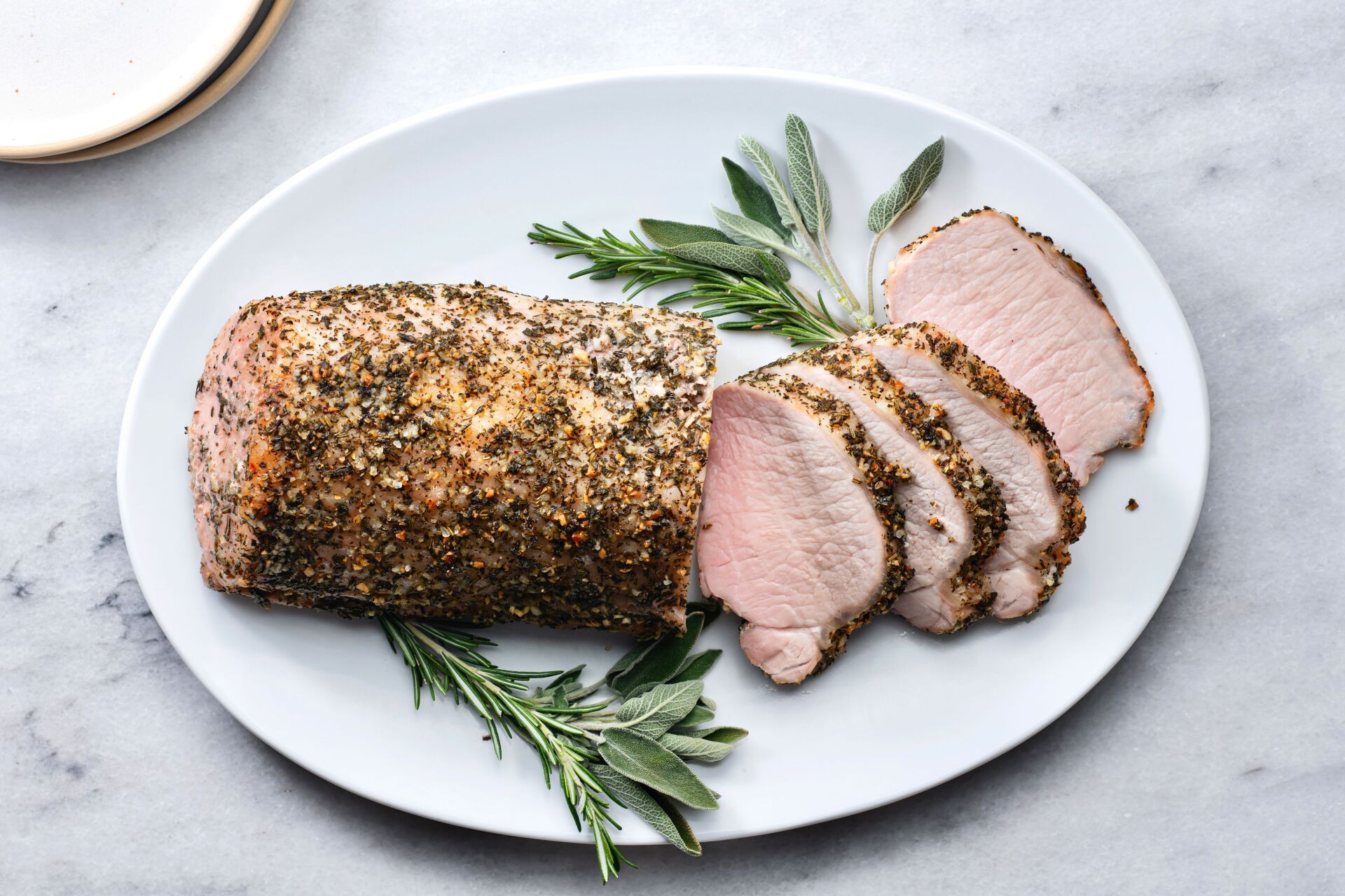 A herb-crusted pork roast, partially sliced, sits on a white oval platter garnished with fresh rosemary and sage sprigs.