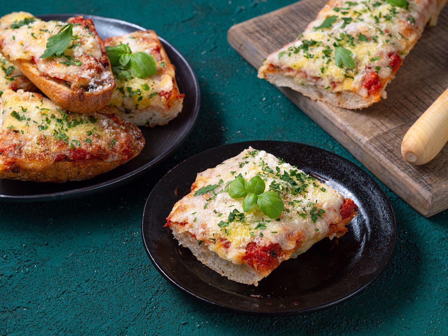 Various portions of baked French bread pizza topped with melted cheese, tomato sauce, and fresh herbs on plates and wood.