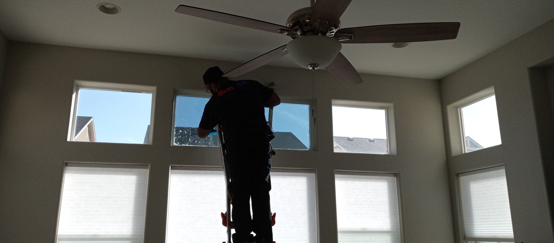A man is standing on a ladder in a room with a ceiling fan and he's installing window tint.