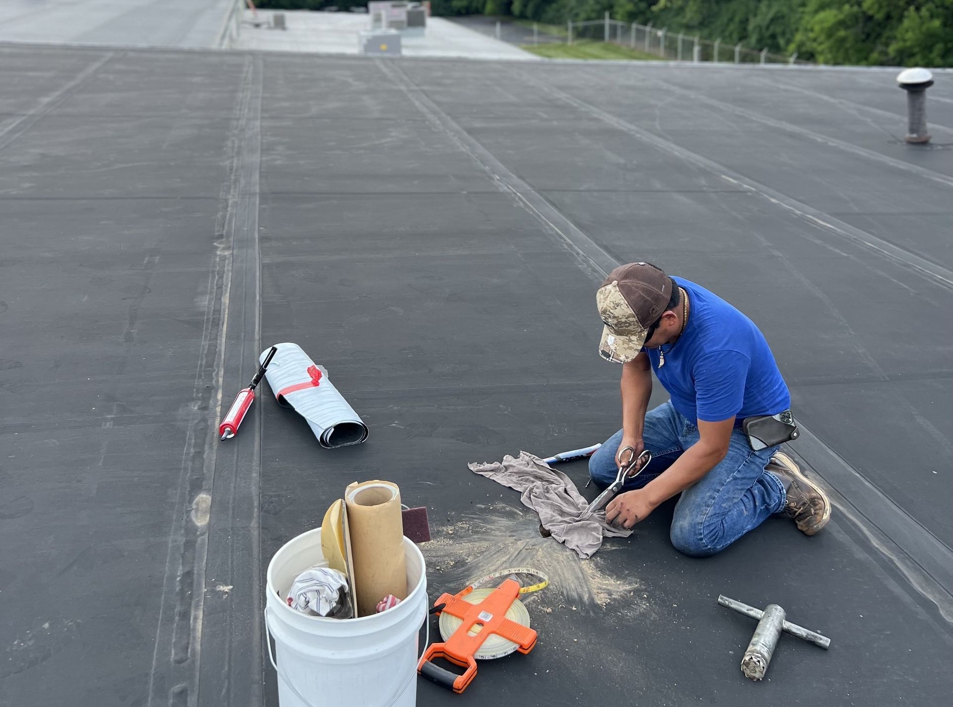 Worker kneeling on a flat roof beside tools and roofing materials, inspecting a seam near a bucket.
