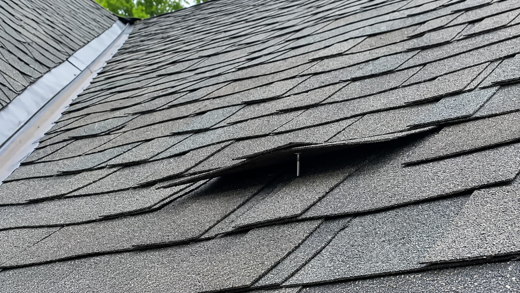 Damaged gray shingle roof with a hole and lifted shingles near the edge