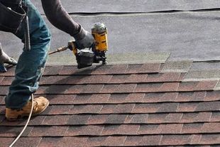 Worker using a nail gun to install brown shingles on a roof