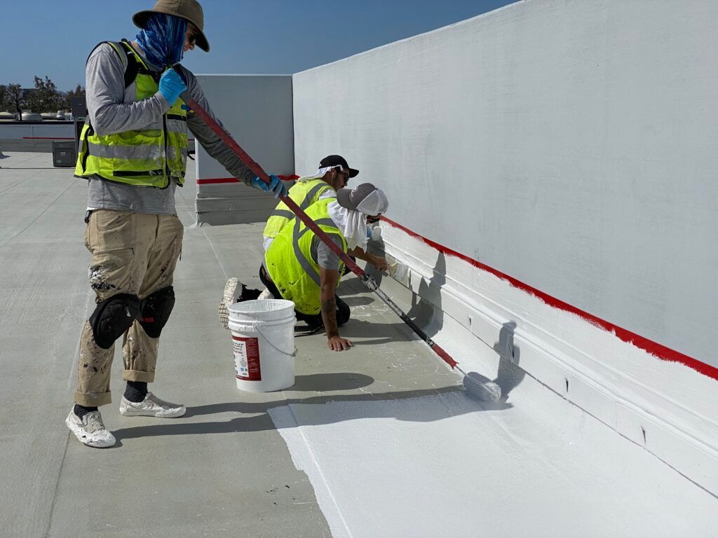 Two workers painting a white wall outdoors, one using a roller and one kneeling near paint buckets.