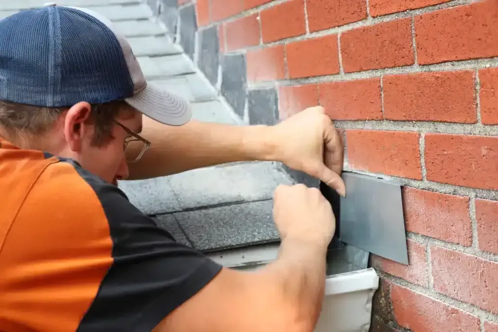 Worker installing gutter on a brick wall beside gray roof shingles, using a hand tool.
