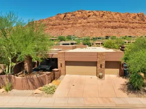 Desert home with tan garage and driveway, framed by green trees and red rock cliffs under a clear blue sky