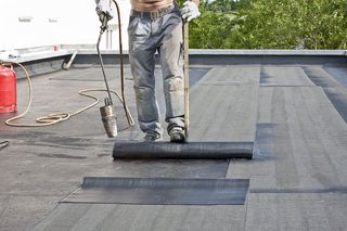 Worker installing black roofing felt on a flat roof with a torch applicator.