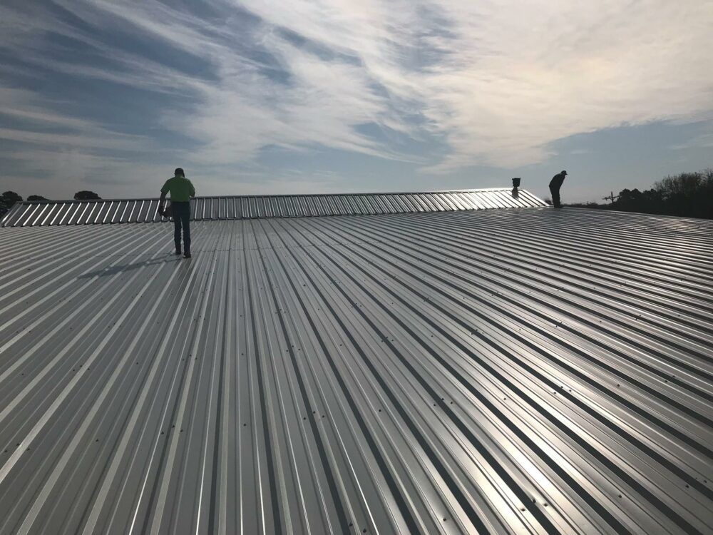 Workers on a large corrugated metal roof under a cloudy sky
