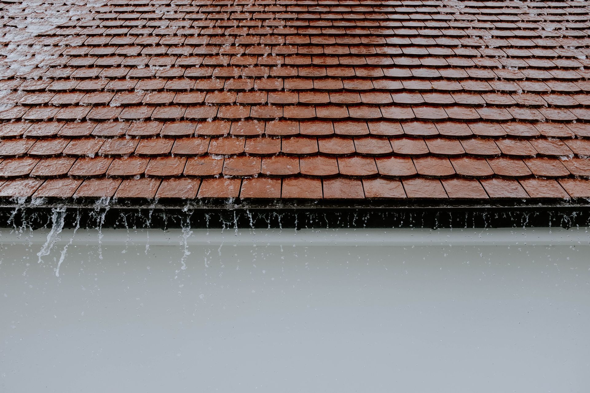 Water spilling off a brown tiled roof above a light gray wall.