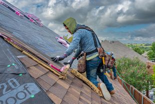 Worker installing shingles on a steep roof under cloudy skies, wearing a hooded jacket and harness.