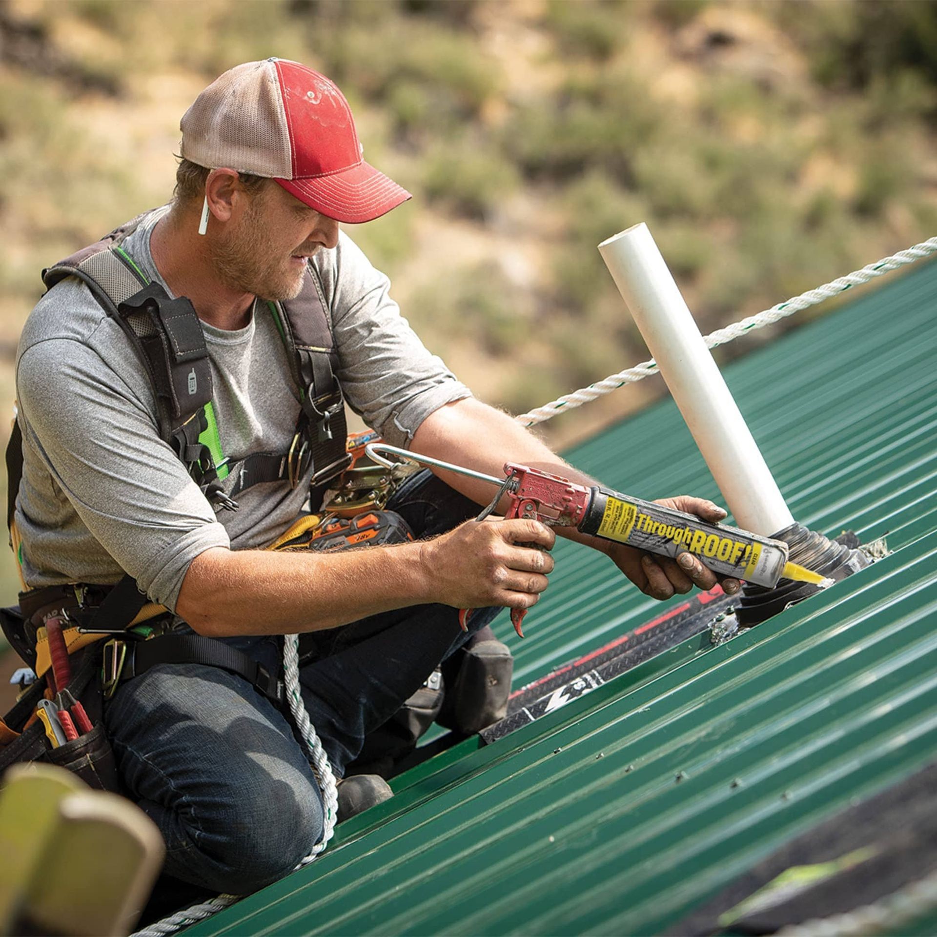 Worker applying sealant to a green corrugated metal roof while kneeling in a harness and cap
