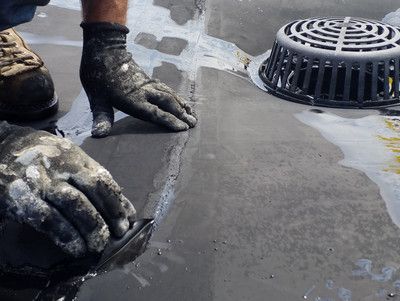 Hands smoothing wet black roofing material around a drain on a flat roof.
