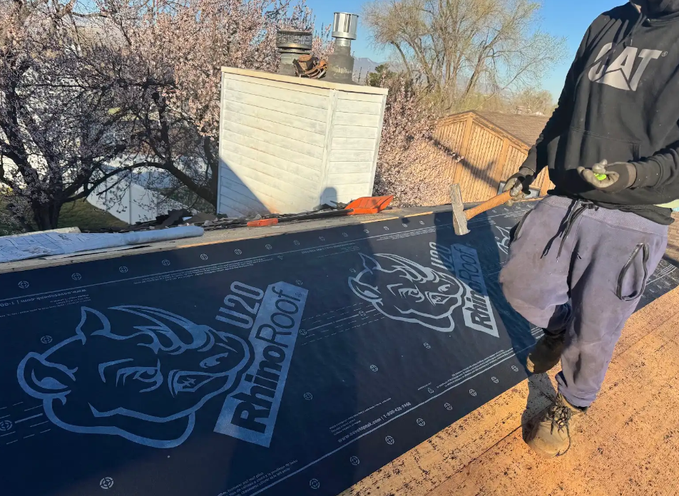 Worker installing black roofing underlayment on a roof, with a white vent and trees in the background.