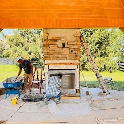 Two men are working on a stone fireplace under a wooden roof.