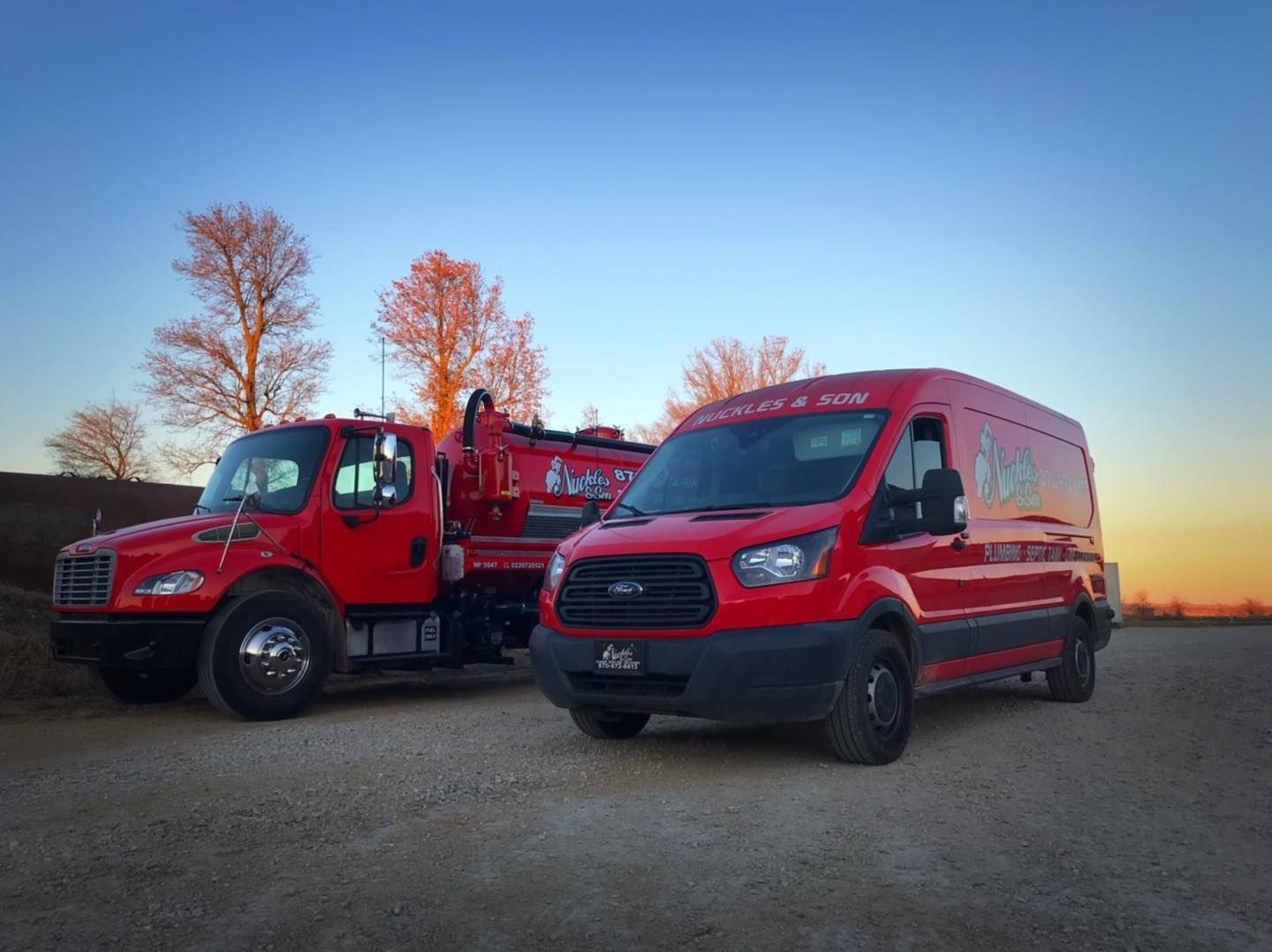 Two red utility vans parked on a gravel lot at sunset with bare trees in the background