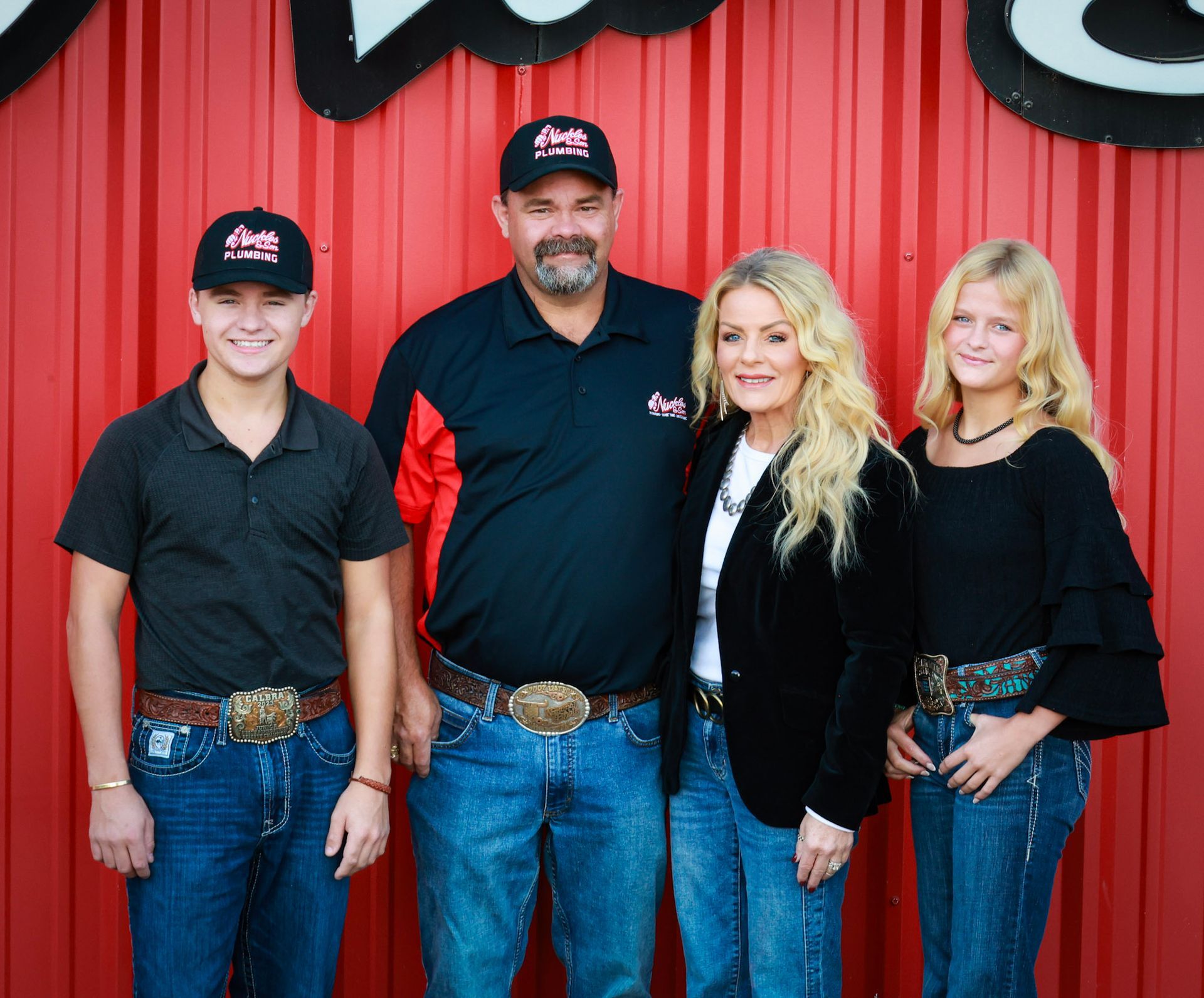 Four people in black shirts and jeans pose in front of a red corrugated wall.