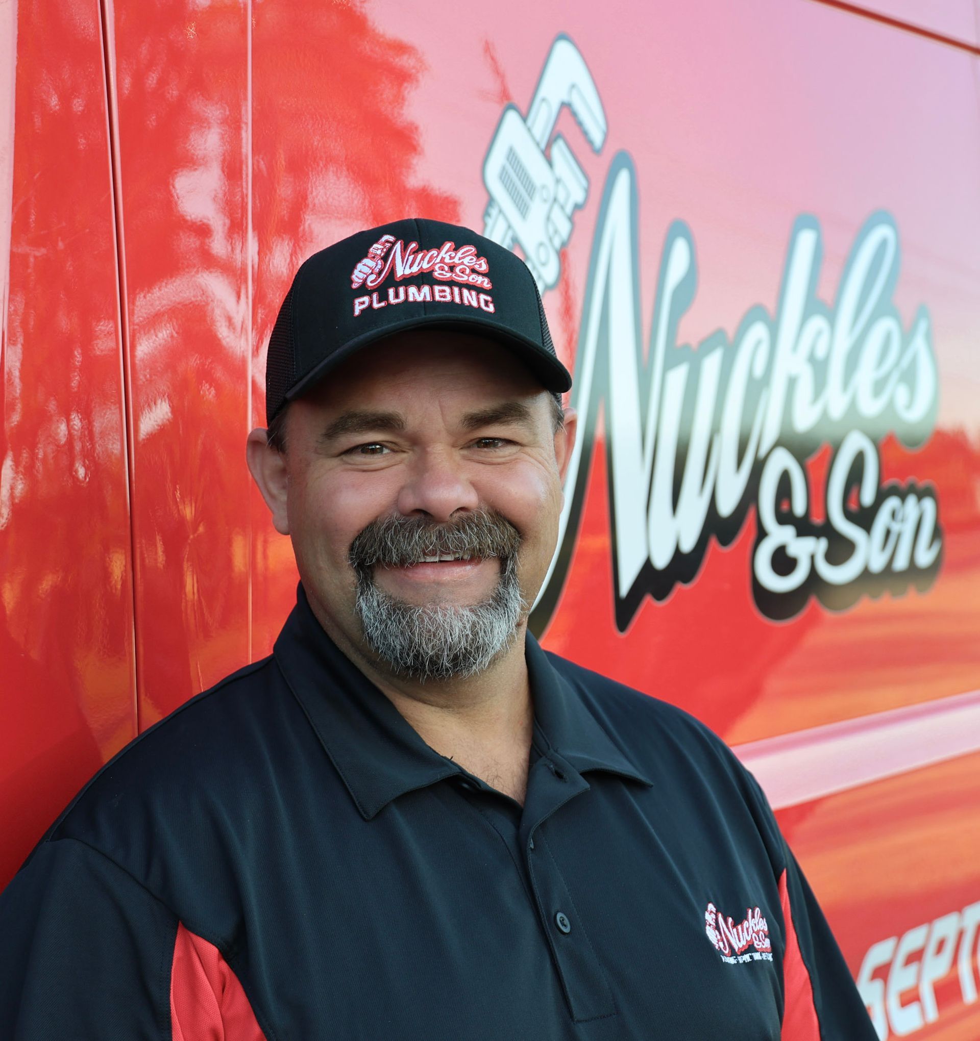 Man standing before a red Muckles & Son truck, smiling in a black cap and polo shirt.
