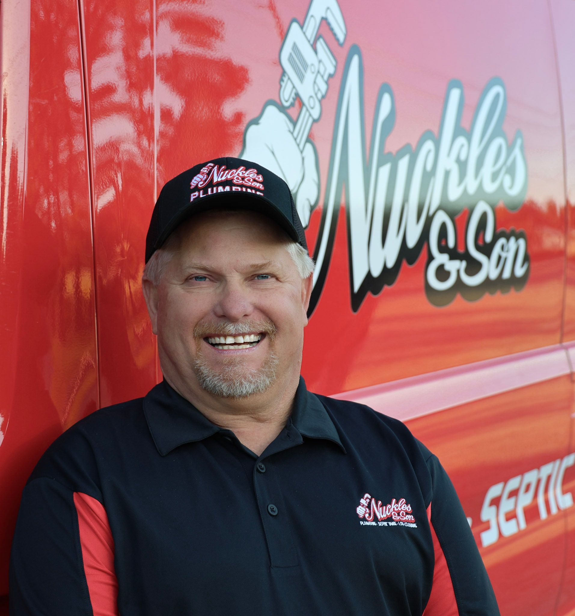 Smiling employee in a black polo and cap standing in front of a red Nickles & Son service truck.