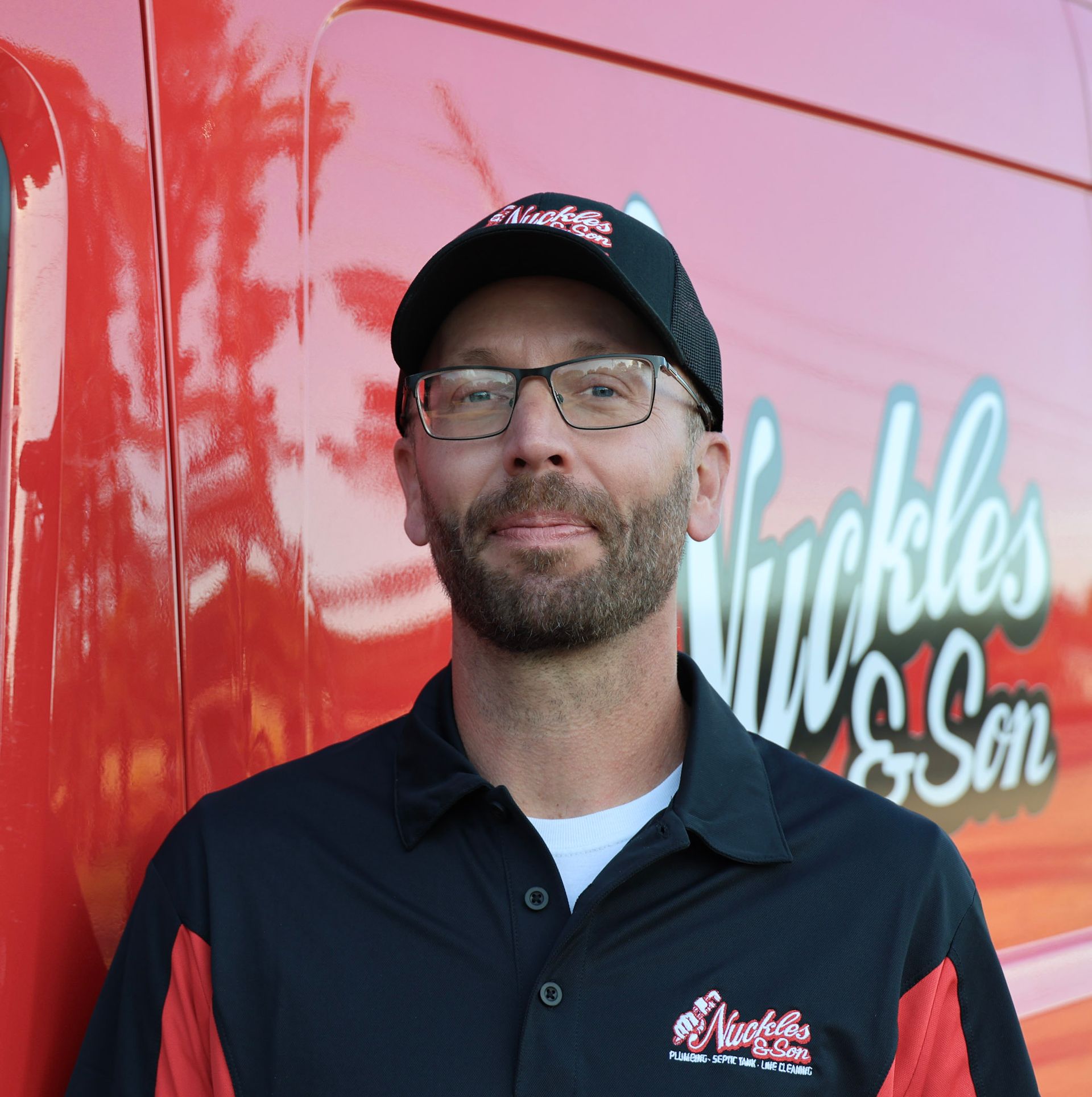 Man in glasses and cap standing before a red truck with “Nickles” logo, smiling gently